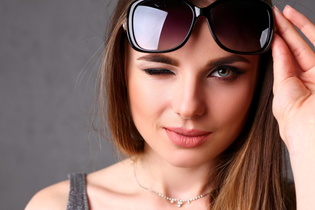 Nahaufnahme einer jungen Frau mit Sonnenbrille, die zwinkert und leicht lächelt.